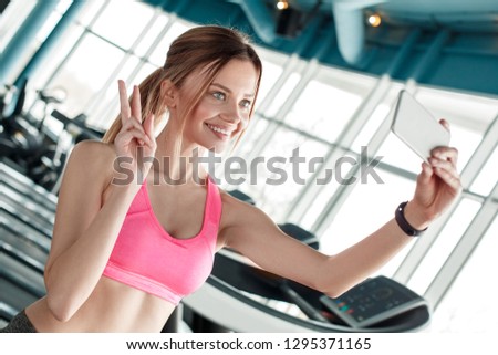 Young woman in gym healthy lifestyle standing holding smartphone taking selfie pictures looking camera smiling cheerful showing salute gestue