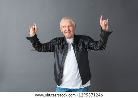 Senior man wearing black leather jacket standing isolated on gray wall hands up showing horns sign looking camera smiling cheerful