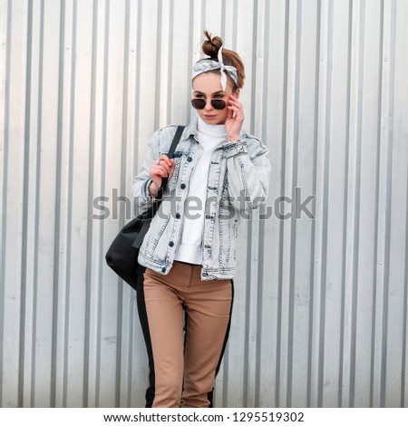 Young hipster girl in trendy clothes in trendy bandana with a stylish hairstyle in sunglasses with a handbag in a knitted sweater near a metal building. American girl. Summer style women's clothing.