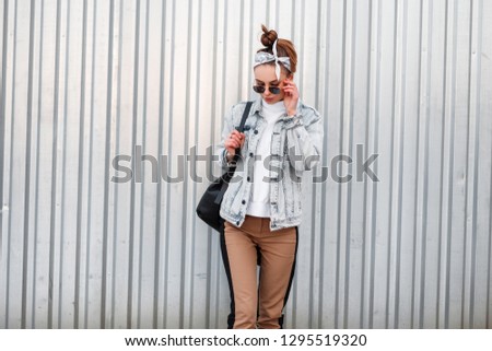 Young attractive hipster woman in knitted vintage white sweater in beige pants in a denim jacket in a bandana with a black backpack posing outdoors near a metal building. Cool girl. Women's fashion. 