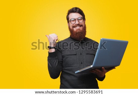 Cheerful bearded man is holding laptop and poiting bkack, looking at camera over yellow background Royalty-Free Stock Photo #1295583691
