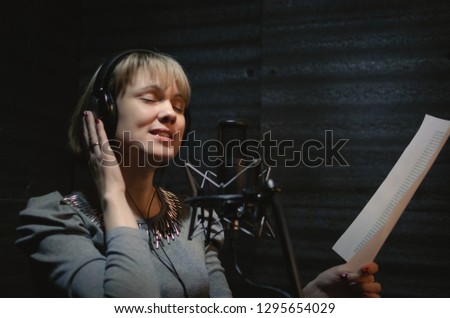 Woman a singer in a headphones is performing a song in a sound recording studio and is standing in front of the microphone.