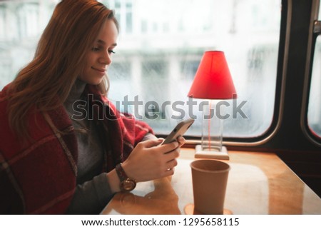 Girl with a cup of coffee sits in a coffee shop and uses a smartphone, a woman covered with a blanket. Attractive girl sits in a retro cafe and looks at the smartphone screen. Cozy portrait in cafe