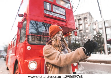 Cute positive girl with winter clothes standing near the red bus and cafe to take selfie on smartphone. Attractive girl walking along the streets of the winter town and take selfie.