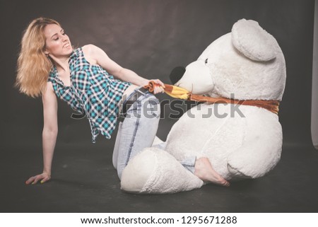 cute blonde teen in jeans and plaid shirt plays with her huge Teddy polar bear in yellow scarf on black background alone