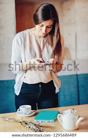 Pretty young girl blogger taking a photo with mobile phone while standing at the table in a cafe