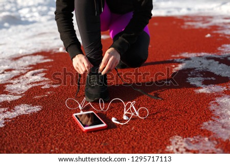 girl athlete in black sneakers, crouching on the red track for running. near is a phone with wired headphones. Cold snowy weather.