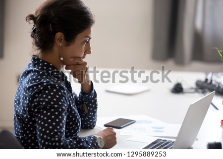 Indian girl sitting at desk near computer cogitating thinking making important decision at workplace. Concentrated serious office worker millennial woman analysing results feels doubts and feel unsure Royalty-Free Stock Photo #1295889253