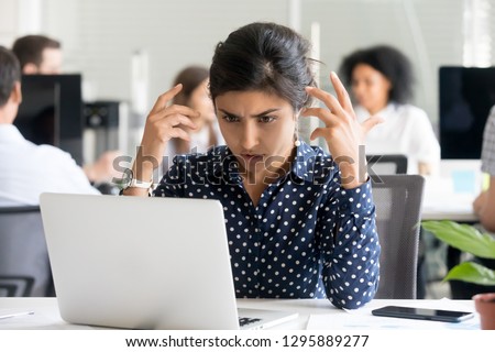 Upset indian female worker sitting at desk in office looking at pc screen feels unhappy and shocked. Millennial hindu woman reading unpleasant notification having system fault and big troubles concept Royalty-Free Stock Photo #1295889277