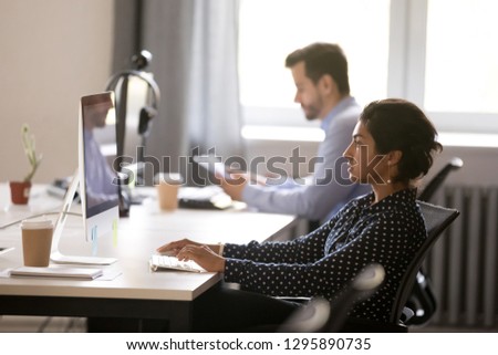 Side view diverse employees sitting at desk using laptop, focus on young indian female. Millennial serious hindu worker typing message business letter on computer looking at screen searching new ideas Royalty-Free Stock Photo #1295890735