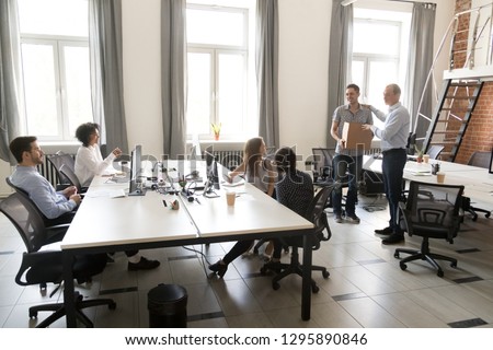 Office workers sitting on their places looking at boss ceo introduce a new employee to colleagues. Millennial man holds box has first workday in company getting vacancy feels satisfied and happy Royalty-Free Stock Photo #1295890846