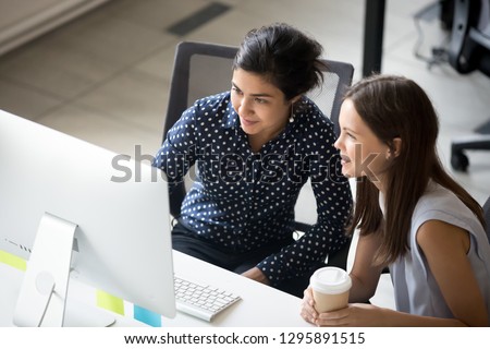 Multiracial colleagues indian and caucasian young women having coffee break sitting together at desk in office. Diverse students interns friends looks at pc screen talking discussing working moments Royalty-Free Stock Photo #1295891515