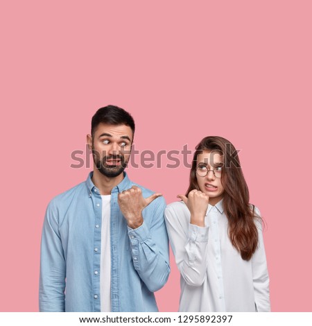 Puzzled two friends point at each other with displeasure, wears shirt, cant decide who should do work, have misunderstanding, isolated over pink background with blank space for your promotion