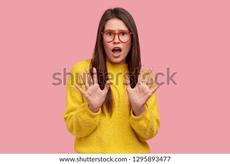 Displeased surprised dark haired young woman demonstrates bad attitude, stop gesture with her palms outward, dressed in yellow casual loose jumper, shows rejection, isolated on pink studio wall