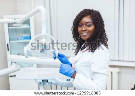Portrait of Female black dentist in dental office. She standing at her office and she has beautiful smile. Modern medical equipment Royalty-Free Stock Photo #1295916085