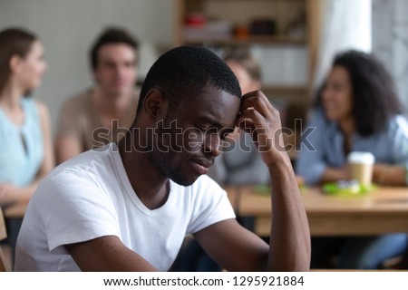 Sad African American man suffering from bullying or racial discrimination, sitting alone in cafe, feeling outsider, gossips, low self-esteem, depressed black guy rejected by Caucasian people Royalty-Free Stock Photo #1295921884