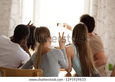 Rear view happy multiracial friends making selfie, recording video, posing for photo for social networks, having fun, spending free time, weekend together in cafe, focused looking at smartphone