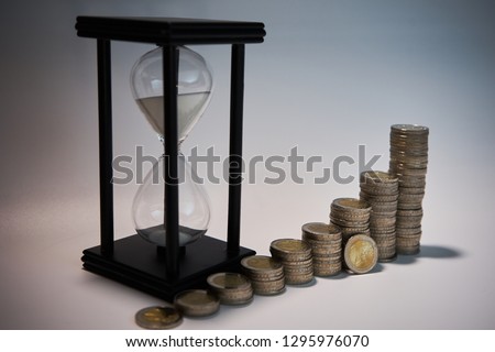 Black sand clock with white sand behind growing staples of coins and a two euro coin leaning against them