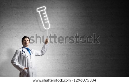 Young confident doctor in white medical uniform interracting with glowing vial symbol whie standing against dark gray wall on background.