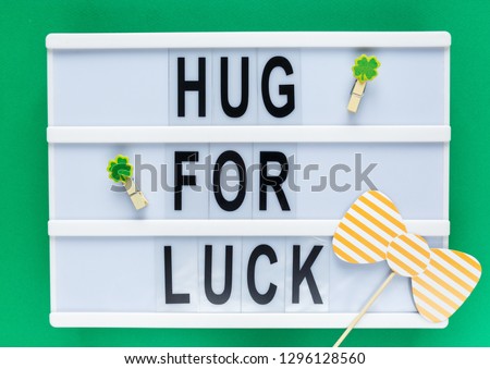 Lightbox with title Hug for luck and photobooth orange bow tie on wooden sticks at green background. Creative background to St. Patricks Day