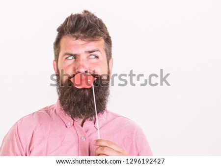 Man with trendy beard holding paper lips. Bearded man making funny faces isolated on white background. Hipster with long beard in pink shirt posing with paper accessories, party memories concept.