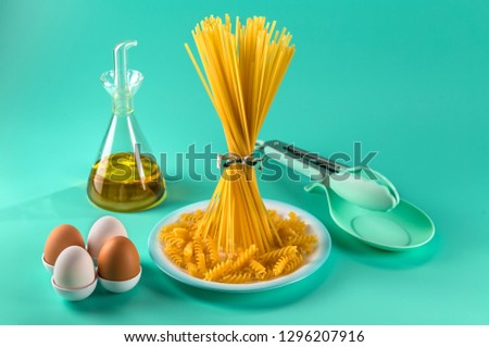 bunch of spaghetti standing upright on a bright colored background surrounded by chicken eggs, olive oil and cooking utensils. Horizontal.