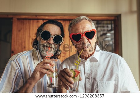Senior men wearing funny sunglasses drinking juice with straw. Elderly friends with crazy eyewear having juice indoors.