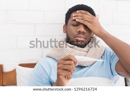 Sick african-american man having fever, measuring body temperature with thermometer, touching forehead in bed, copy space Royalty-Free Stock Photo #1296216724
