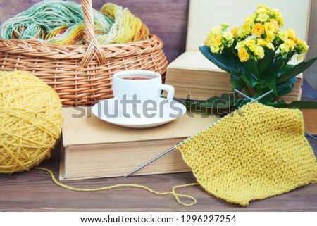 Knitting of yellow wool with needles, white espresso coffee cup saucer, basket, old books, Kalanchoe on wooden rustic background. Front view