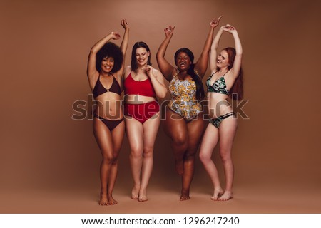 Full length of different size women in bikinis dancing together over brown background. Multi-ethnic women in swimwear enjoying themselves.