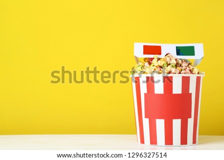 Sweet popcorn in striped bucket with glasses on yellow background