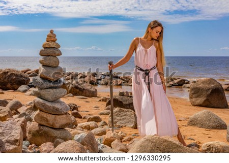 Young woman in long dress, in hands holds a Viking sword.Model posing on background of sea shore and zen pyramid of stones