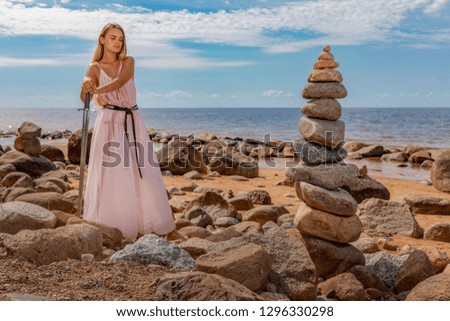 Young woman in long dress, in hands holds a Viking sword.Model posing on background of sea shore and zen pyramid of stones
