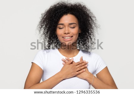 Grateful hopeful happy black woman holding hands on chest feeling pleased thankful, sincere african lady expressing heartfelt love appreciation gratitude honesty isolated on white studio background Royalty-Free Stock Photo #1296335536