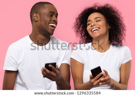 Happy young african american couple laughing using smartphones apps isolated on pink studio background, cheerful black friends users holding mobile phones gadgets having fun with cellphones together  Royalty-Free Stock Photo #1296335584