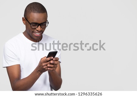 Smiling black young man in glasses holding phone looking at smartphone isolated on white grey studio background with copy space aside, happy african guy using mobile applications texting on cellphone
