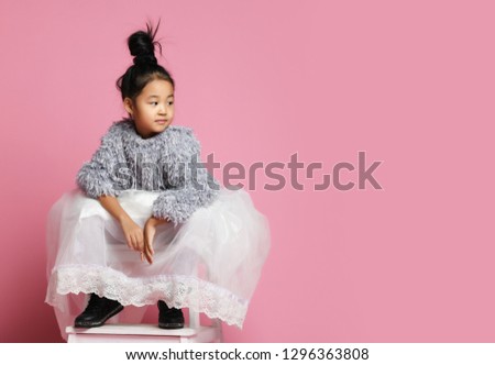 Young asian girl kid in long white skirt, grey fluffy sweater and black shoes sitting and looking the direction the text. On pink background. With free text space