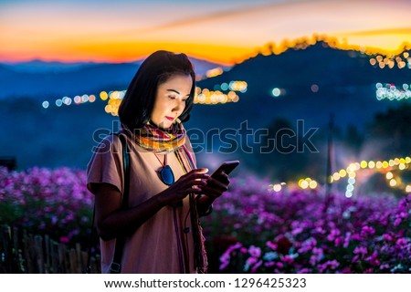 Asian Woman Standing Among Flowers Meadow and Chatting Online Mobile Phone with Beautiful Sunset Bokeh Background at Mon Jam, Chiang Mai, Thailand.