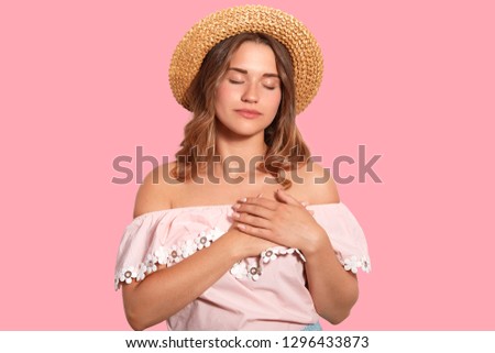 Calm relaxed woman keeps hands on heart, expresses gratitude, being touched or impressed by heart piercing story, wears fashionable summer clothes, isolated over pink background. Feeling concept