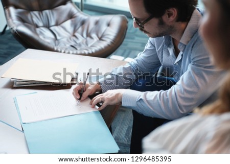 Business man at work in corporate office. Busy hispanic manager with eyeglasses working and meeting with young assistant. People sitting in waiting room and signing documents.