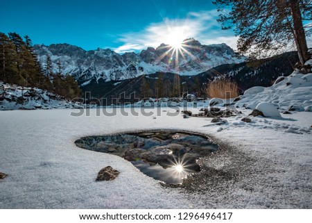 The wonderful Eibsee (lake Eibsee) in Bavaria, Germany, with ice and snow - the mountain Zugspitze is in the back with clouds and the sun