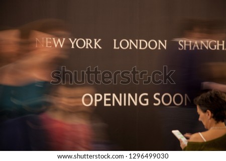 Billboard announcing the opening of a shop as people walk by and a lady sits in front, reading a book.