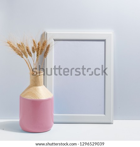 Mock up white frame and spikelets of wheat in pink vase on book shelf or desk. Minimalistic concept.