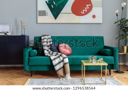 Luxury and stylish interior with green velvet design sofa, coffee table, lamp and commode. Grey walls with abstract painting. Stylish decor of sitting room. Brown wooden parquet.
