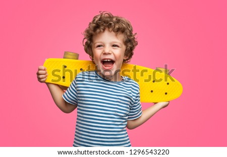 Happy curly boy laughing and holding bright colored skateboard over vibrant pink background isolated