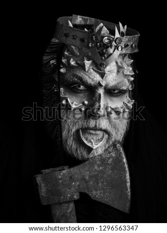 Man with axe on black background. Monster face with white eyes, thorns and warts. Demon with crown on head. King or evil creature with dragon skin and beard. Horror and death concept.