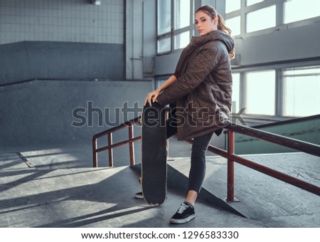 A beautiful young girl wearing a coat with her skateboard sitting on a grind rail in skatepark indoors.