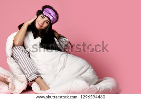 Beautiful brunette girl just woke up with soft blanket and sleepy mask bandage for sleep on pink background