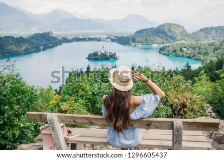 young beautiful girl in a blue dress and a hat sits on a bench on the background of Lake Bled in Slovenia Royalty-Free Stock Photo #1296605437