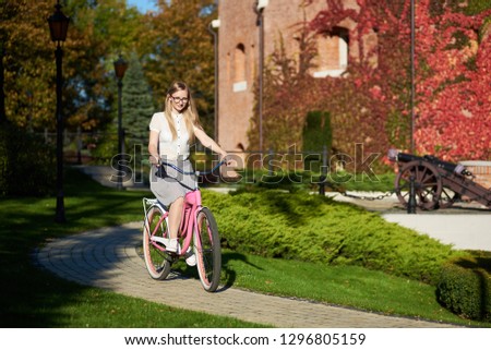 Active blond long-haired attractive tourist woman in glasses and casual clothing cycling modern pink lady bike on bokeh background of beautiful different green trees in the fall.